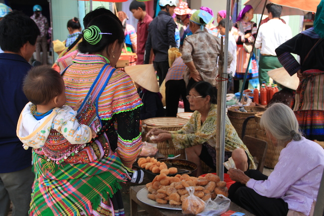 Esta madre demuestra que la riqueza de colores de los vestidos hmong flor incluyen los complementos para llevar a su hijo